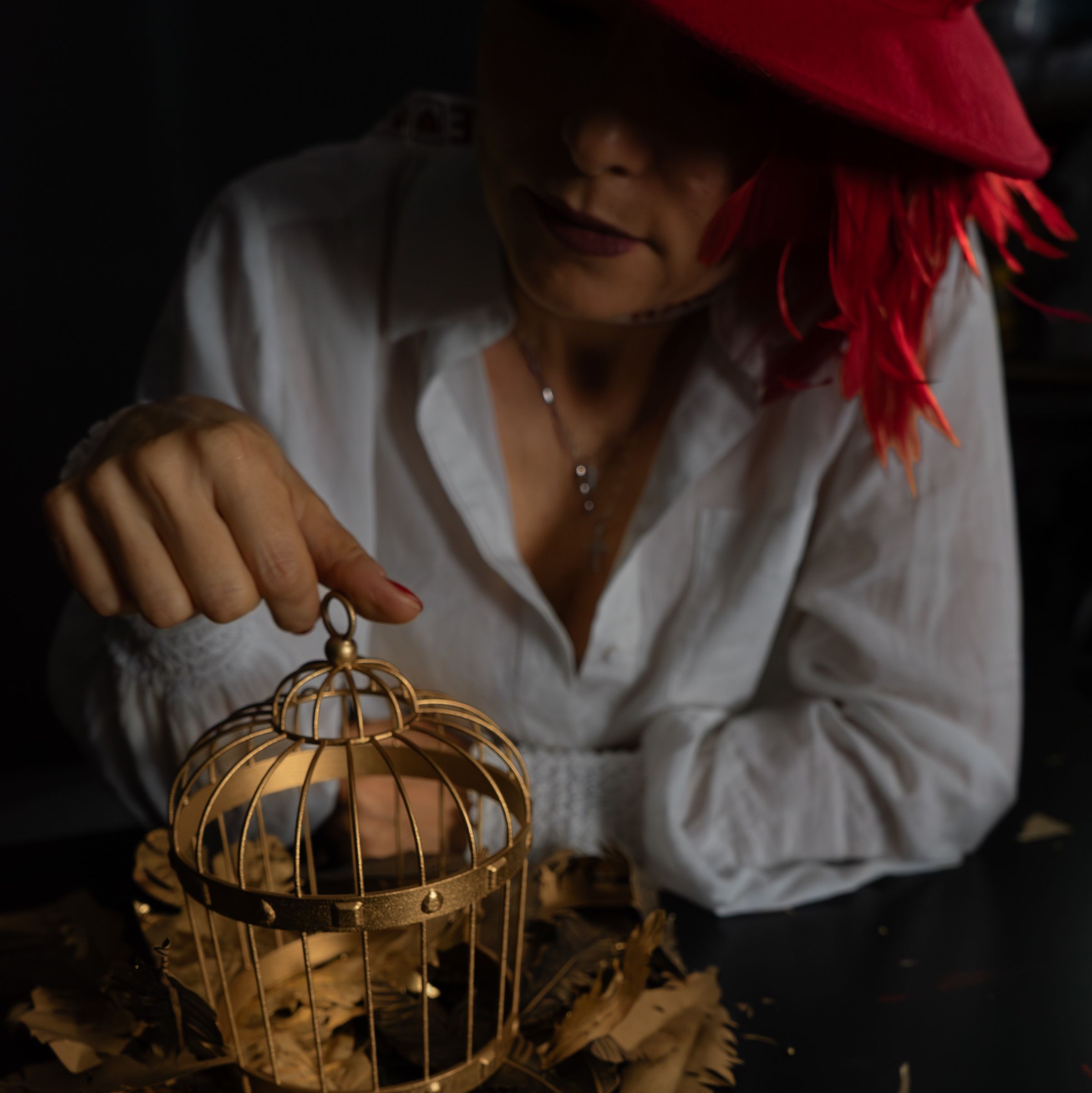 A woman with red feathers in her hair holds a small golden cage in her hands.